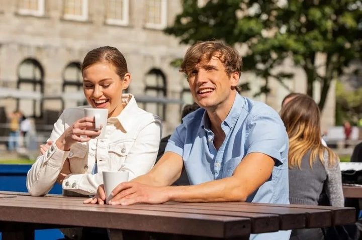 two people sitting on a bench and drinking coffee in the sun