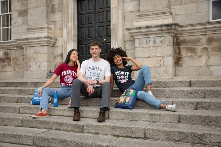 three tourists wearing trinity college merchandise on the steps of trinity college